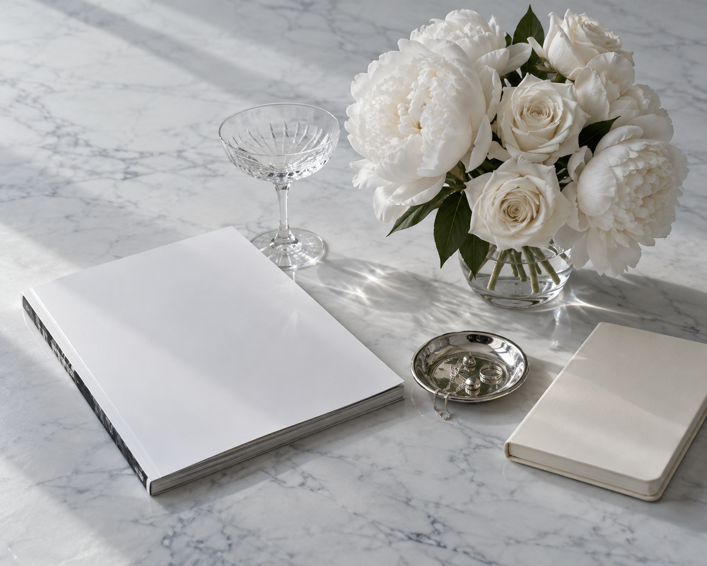 White peonies and roses, a crystal coupe, and a silver jewelry dish in soft daylight